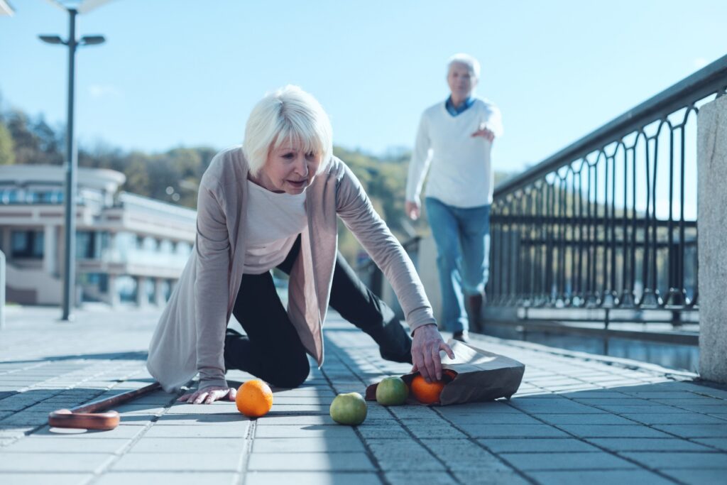 Senior lady fallen on one knee trying to pick up her groceries after tripping on sidewalk
